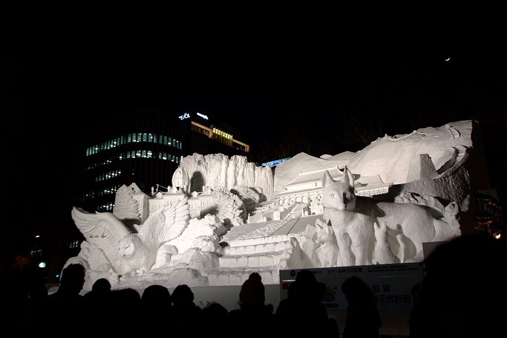 A large illuminated snow sculpture at the Sapporo Snow Festival featuring animals and buildings carved in detailed relief, with visitors standing in front at night.