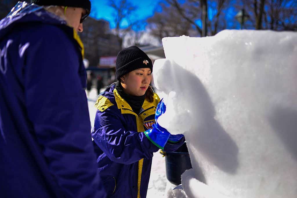 A young woman sculpting snow during the International Snow Sculpture Contest, shaping details into a large snow block while wearing winter gear.