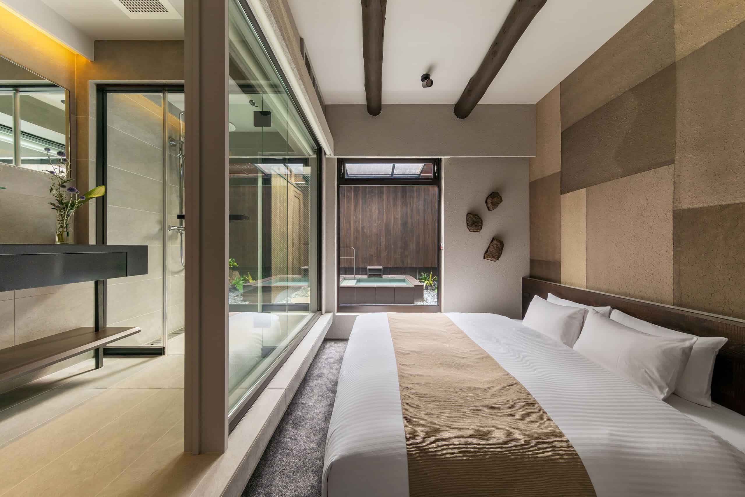 Nazuna Kyoto Tsubaki St. features a cozy bedroom with a glass wall revealing a private open-air onsen bath framed by calming wooden décor.