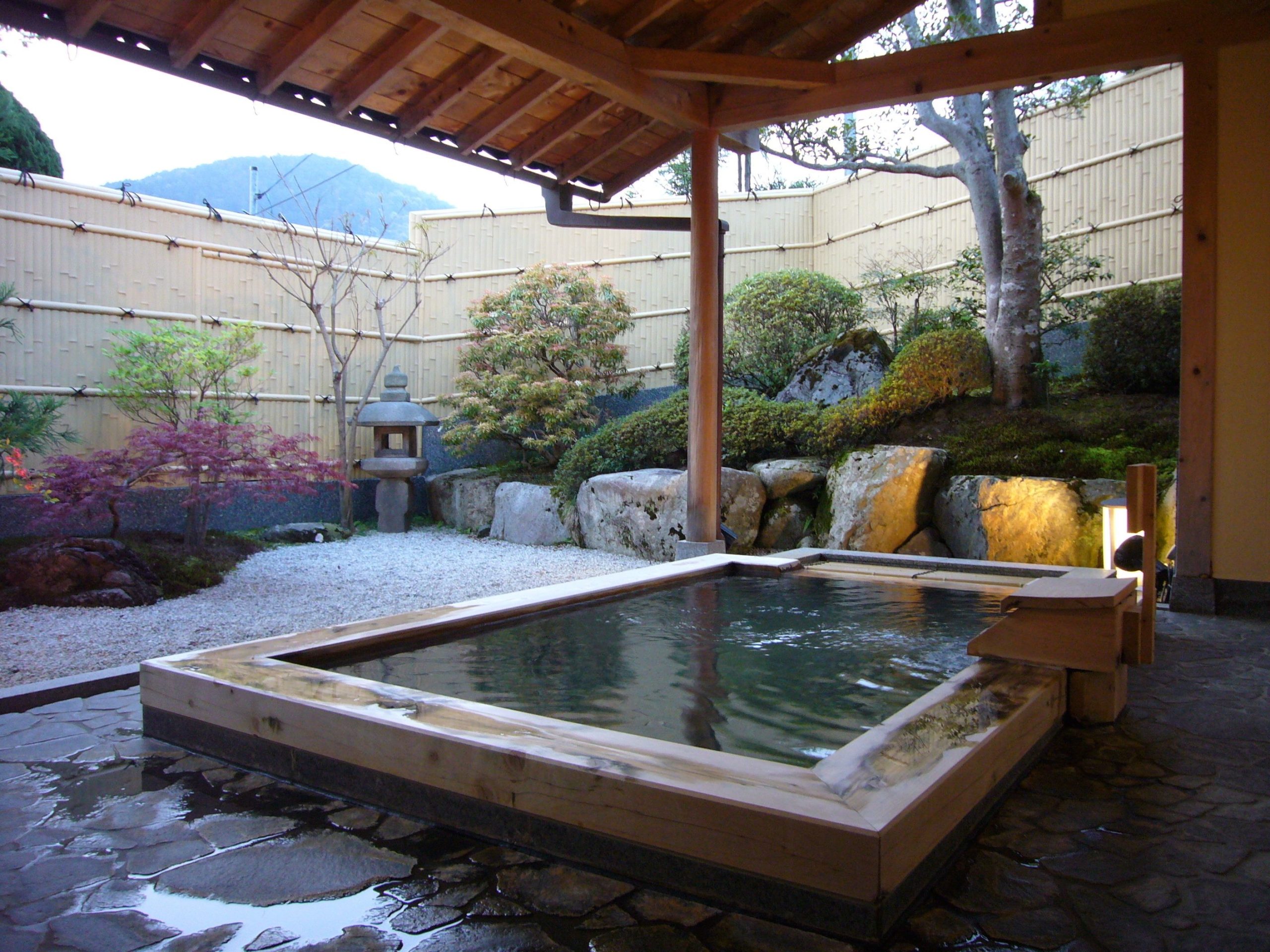 Outdoor onsen bath at Seryo Ryokan in Kyoto, featuring a wooden soaking tub surrounded by a tranquil Japanese garden with stone lanterns, manicured trees, and mountain views.