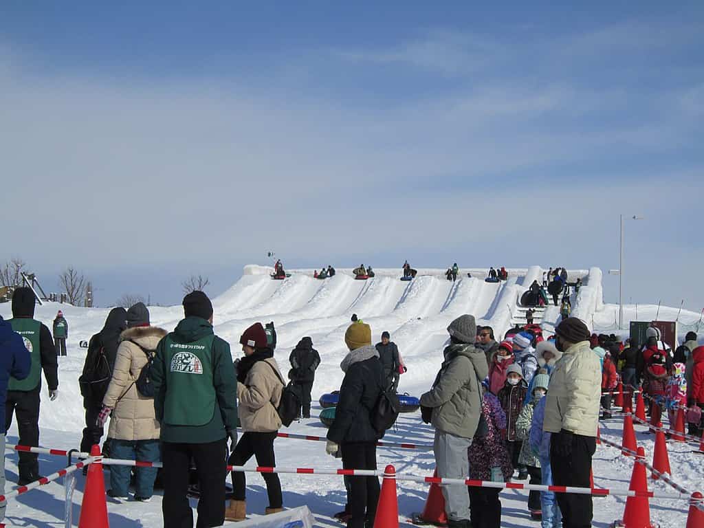 Families and children waiting in line at the Tsudome site with several people tubing down large snow slides under a bright winter sky.