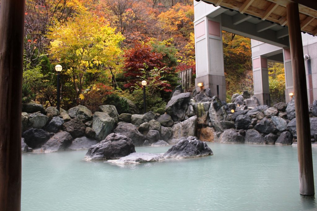 Outdoor hot spring bath at Hotel Mahoroba surrounded by vibrant autumn foliage and natural rock formations in Noboribetsu.