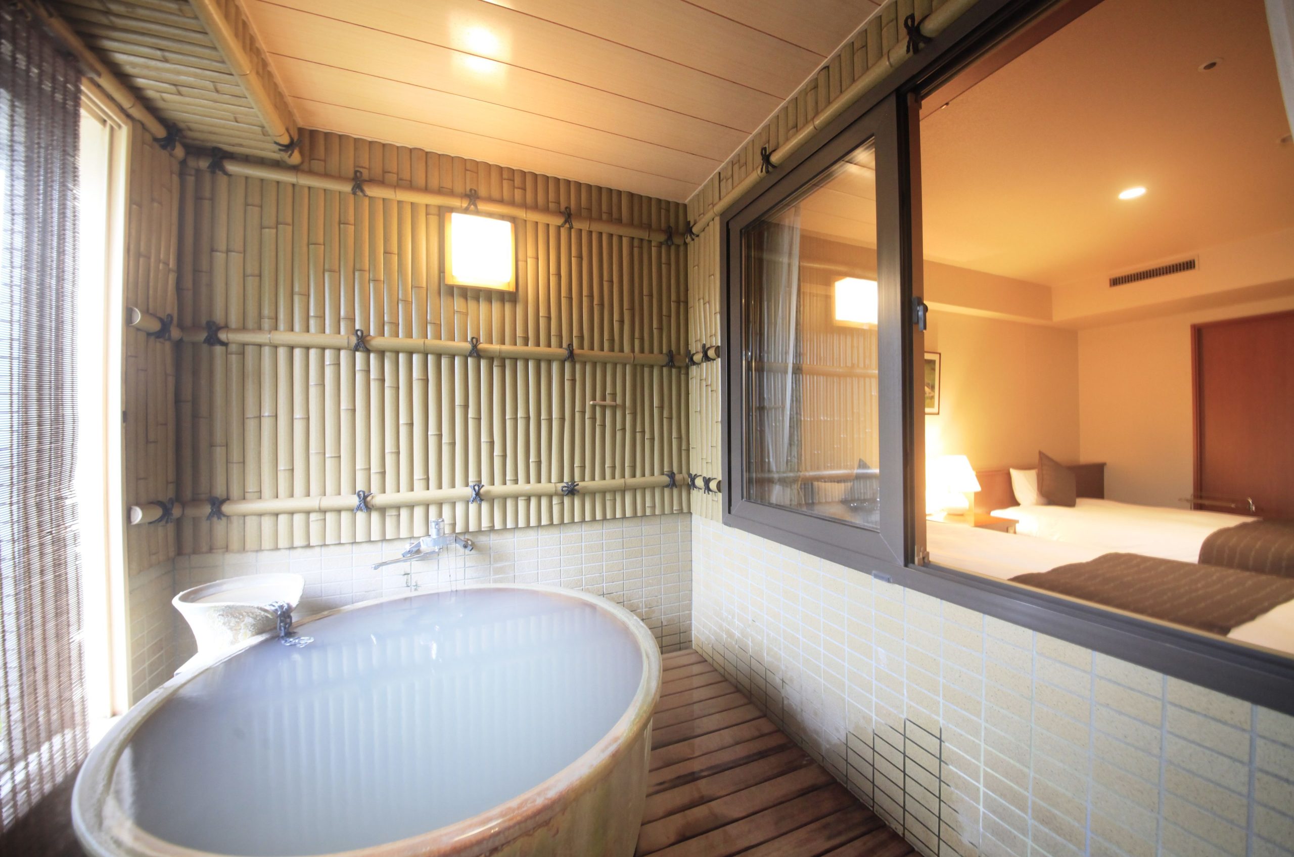 Private milky-white hot spring bath in a bamboo-lined room at Hotel Mahoroba, with a window overlooking the modern guest bedroom