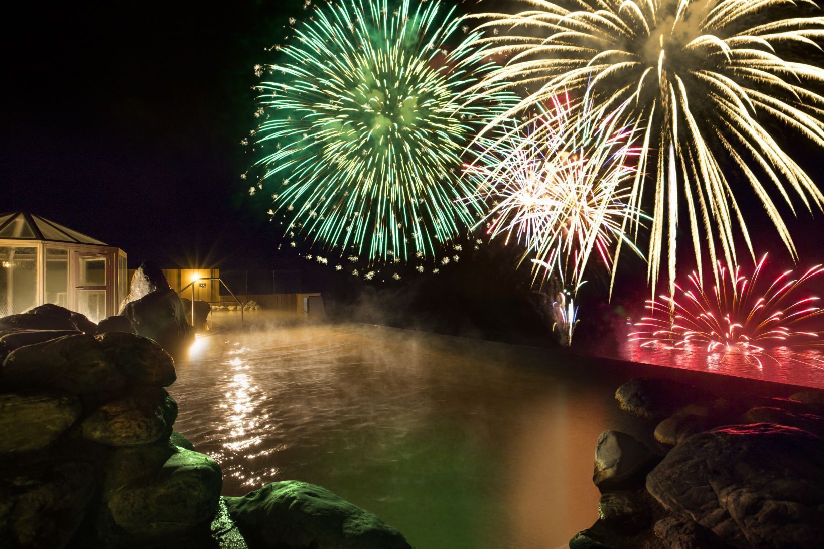 Outdoor hot spring bath at Lake View Toya Nonokaze Resort with vibrant fireworks lighting up the night sky over Lake Toya.