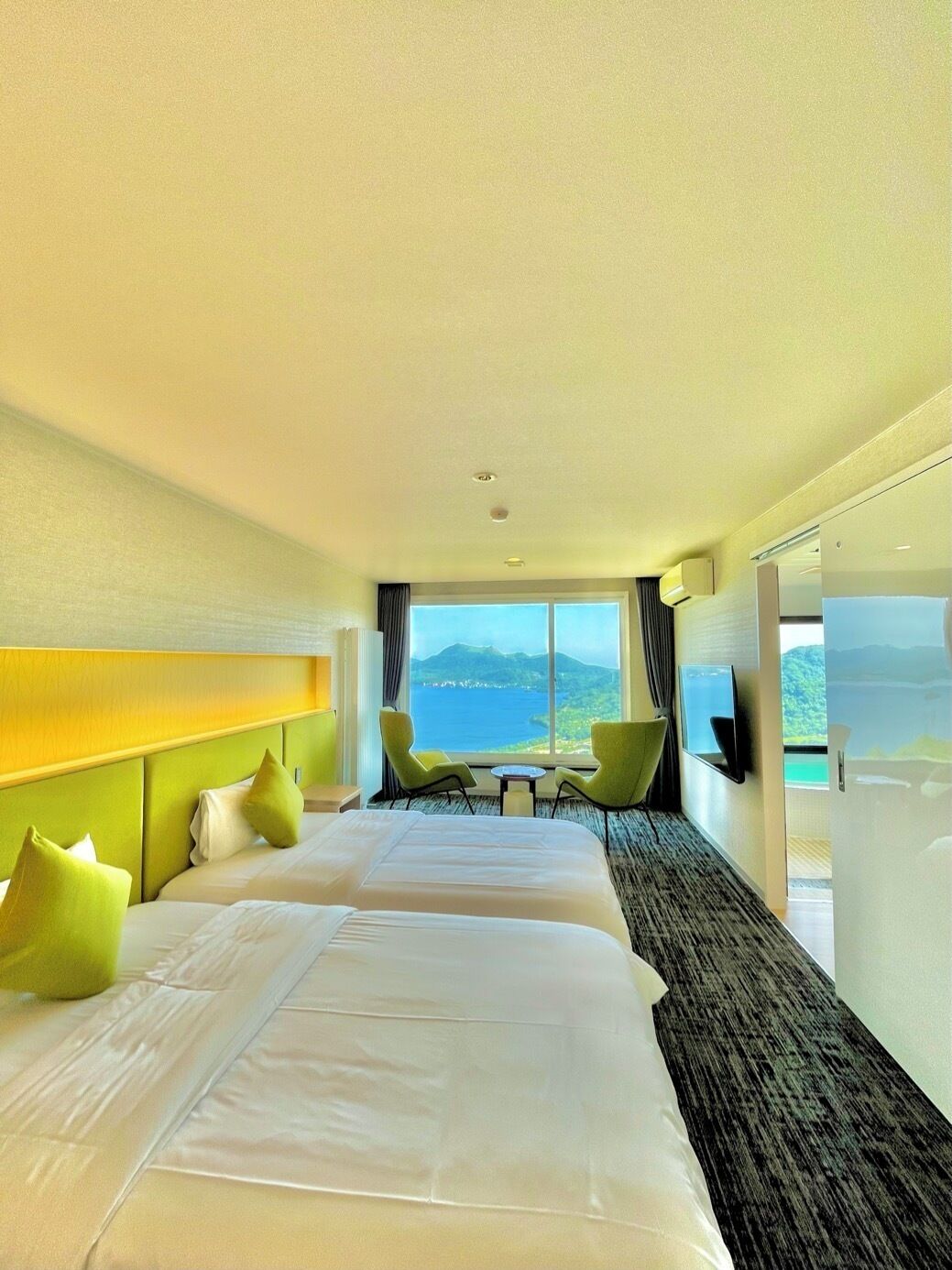 Bright modern guest room at Hotel Cocoa with twin beds, lime-green accents, and a panoramic window overlooking Lake Toya and the surrounding mountains.