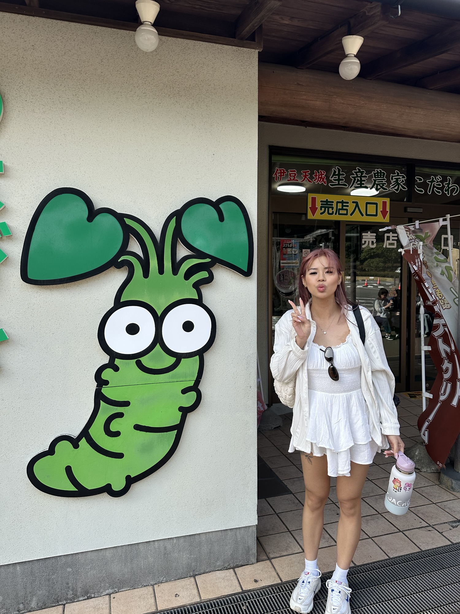 Audrey in a white dress and pink hair standing next to a cartoon wasabi sign in the Izu Peninsula.