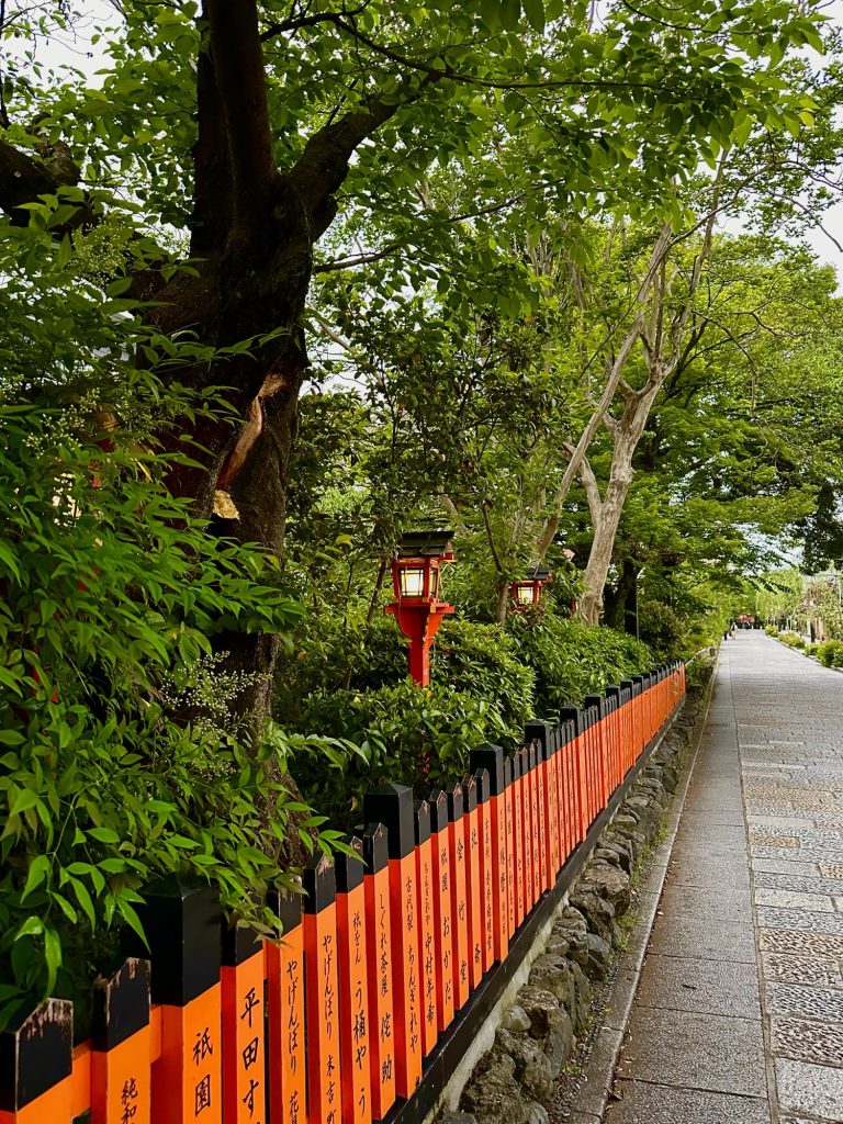 Tree lined pathway beside a red and black fence with lanterns during an evening walk in Gion