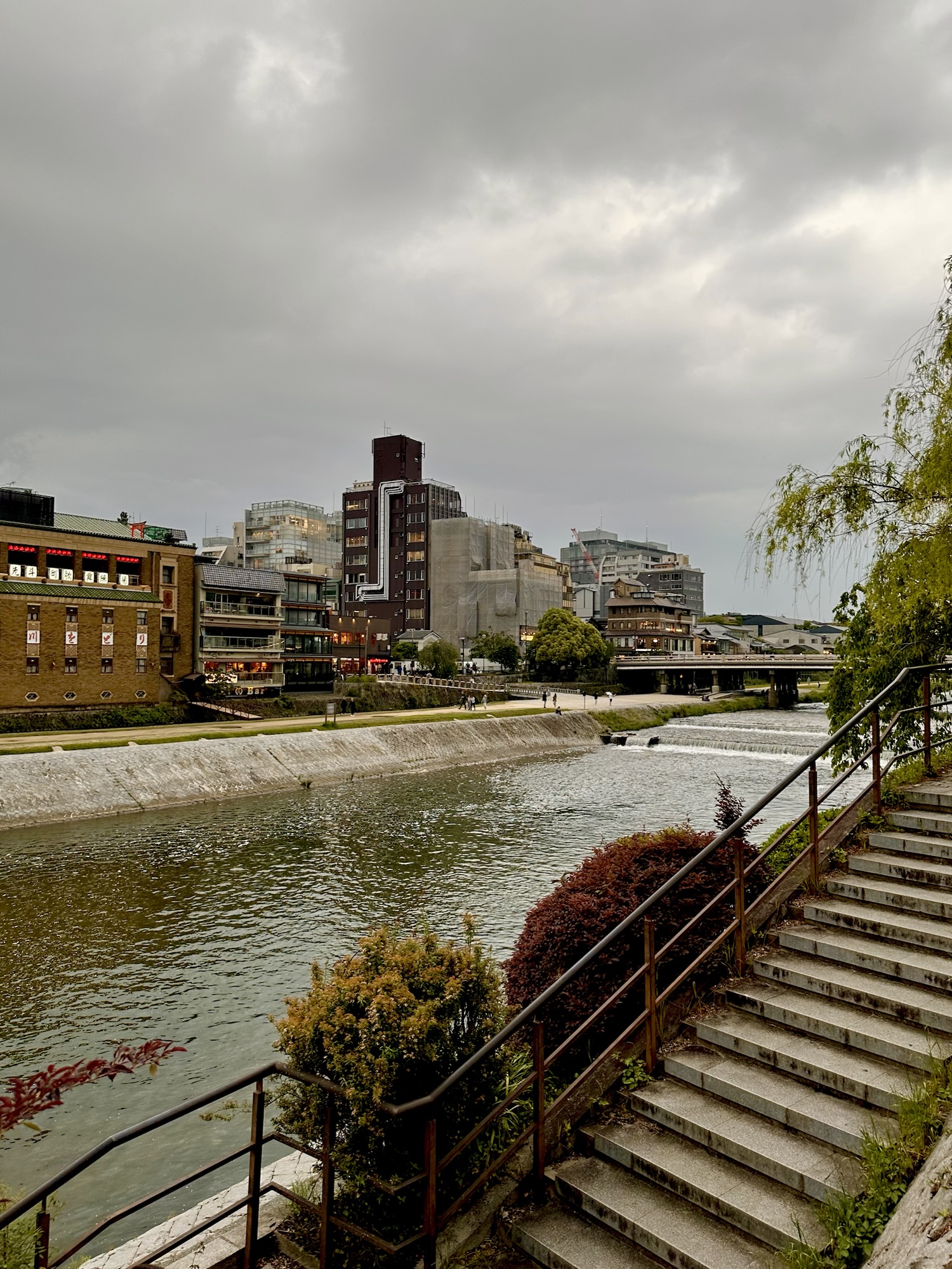View of the Kamogawa River with steps leading down to the water and city buildings under a cloudy sky